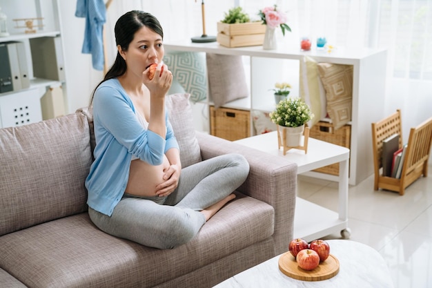 Young pregnant asian woman holding red apple with plate on table in living room. chinese lady mom eats healthy vegan food sitting on couch at home. hungry female pregnancy having energy eating.