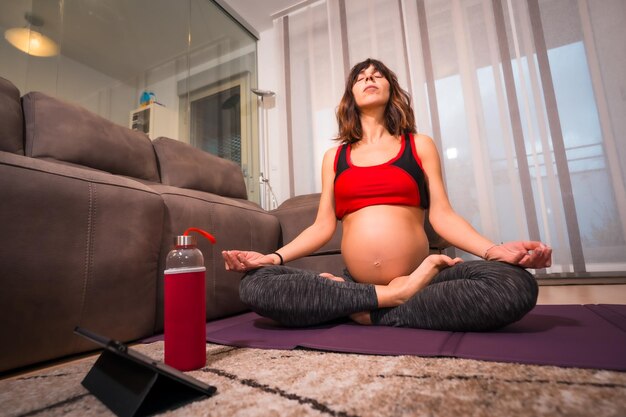 Young pregnant caucasian brunette doing yoga relaxation exercises at home with teacher instructions Online exercises in prepartum classes wellness during pregnancy