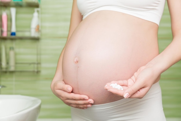 Young pregnant woman applying antistretch marks cream to the abdomen in the morning in the bathroom Pregnancy maternity and bodycare concept