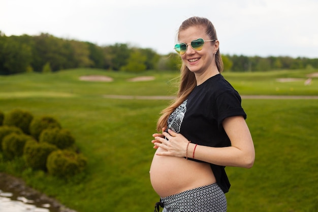 Young pregnant woman in black Tshirt and trousers on the background of green lawn and forest