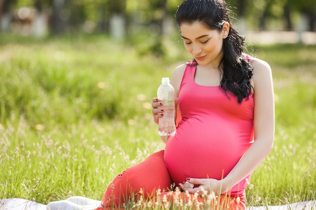 Young pregnant woman doing fitness exercises outdoors and drinking clean bottled water.