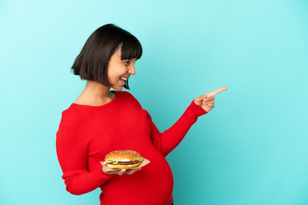 Young pregnant woman holding a burger over isolated background pointing finger to the side and presenting a product