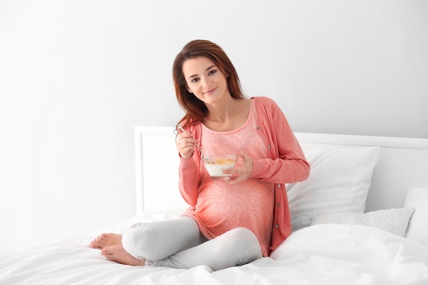 Young pregnant woman holding glass bowl with breakfast and sitting on bed in the room