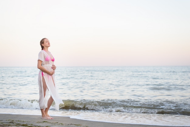 Young pregnant woman is standing on the seashore. Hugging her belly and closed eyes. Enjoying the moment.