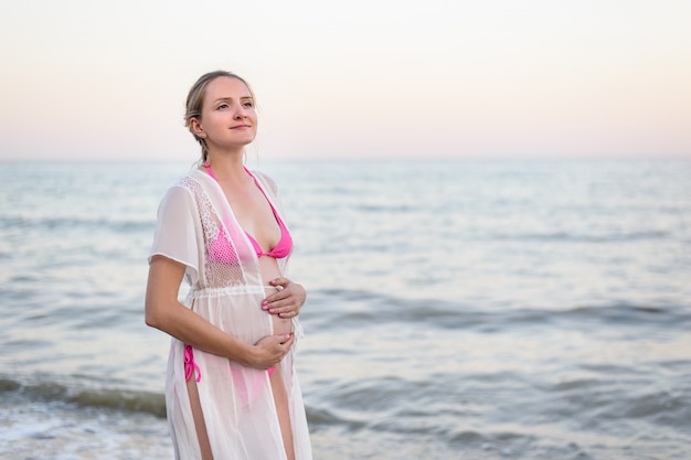 Young pregnant woman is standing on the seashore and hugging her belly. Enjoying the moment.