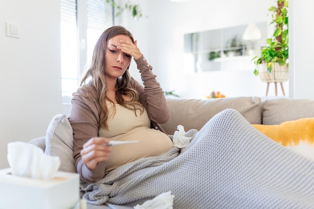 Young pregnant woman lying in bed measuring body temperature holding thermometer feeling sick