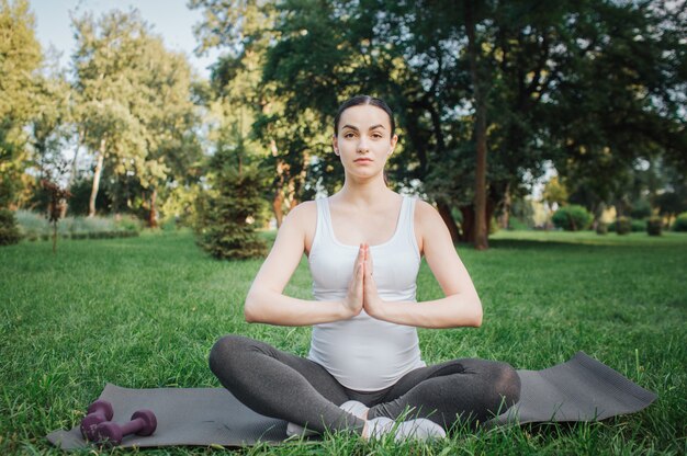 Young pregnant woman meditating outside. She sit on yoga mate in lotus pose. Model keep hands in praying position.