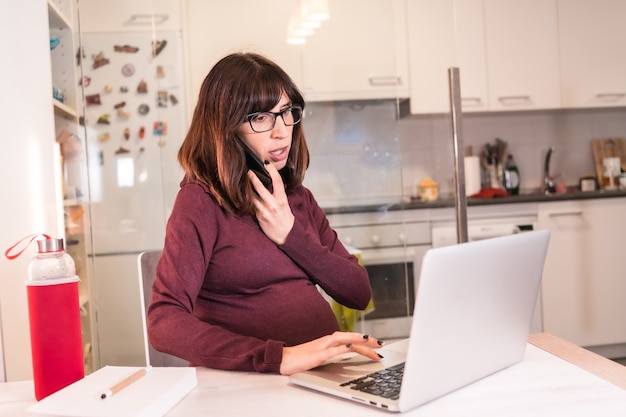 Young pregnant woman teleworking with the computer from home due to the difficulties of working, making a work call