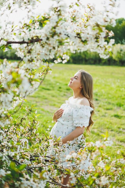 A young pregnant woman in a white dress walks in a blossoming apple orchard Happy pregnant woman Happy future mother