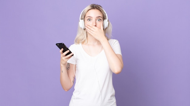 Young pretty albino woman covering mouth with hands with a shocked with headphones and smartphone