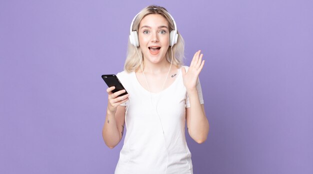 Young pretty albino woman feeling happy and astonished at something unbelievable with headphones and smartphone