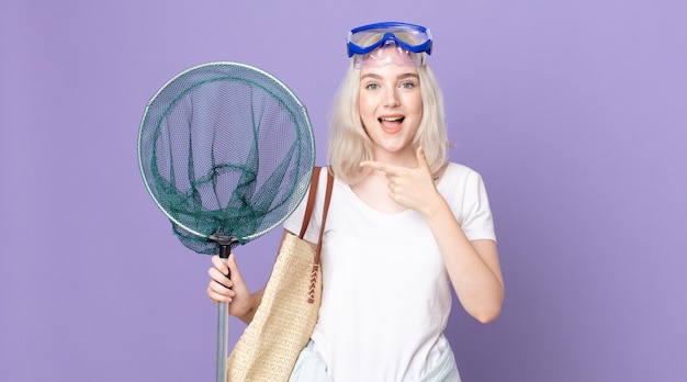 Young pretty albino woman looking excited and surprised pointing to the side with goggles and a fishing net