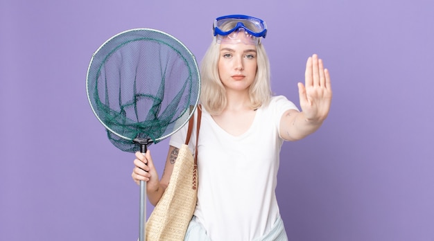 Young pretty albino woman looking serious showing open palm making stop gesture with goggles and a fishing net