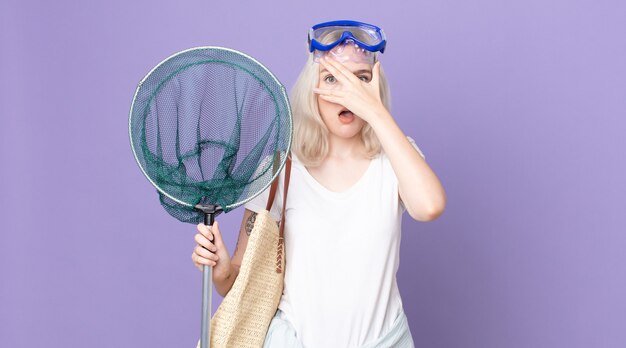 Young pretty albino woman looking shocked, scared or terrified, covering face with hand with goggles and a fishing net