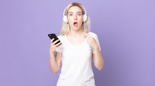 Young pretty albino woman looking shocked and surprised with mouth wide open, pointing to self with headphones and smartphone