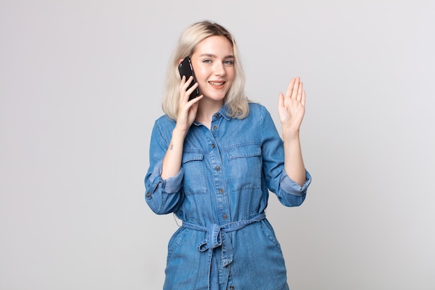 Young pretty albino woman smiling happily, waving hand, welcoming and greeting you and talking with a smartphone