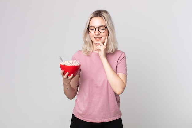 Young pretty albino woman smiling with a happy, confident expression with hand on chin with a breakfast bowl