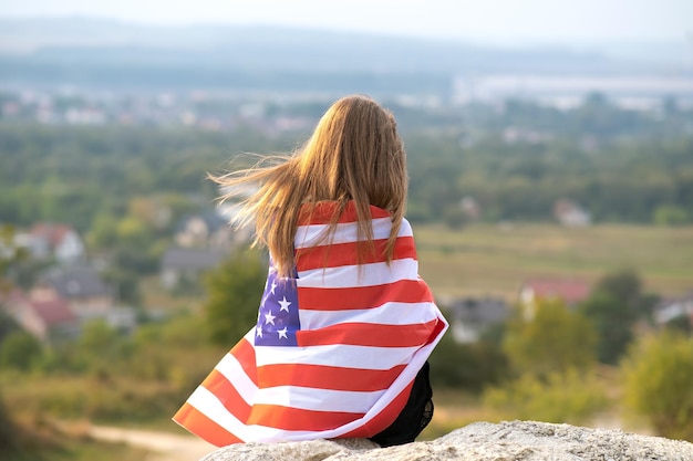 Young pretty american woman with long hair holding waving on wind USA flag on her sholders resting outdoors enjoying warm summer day