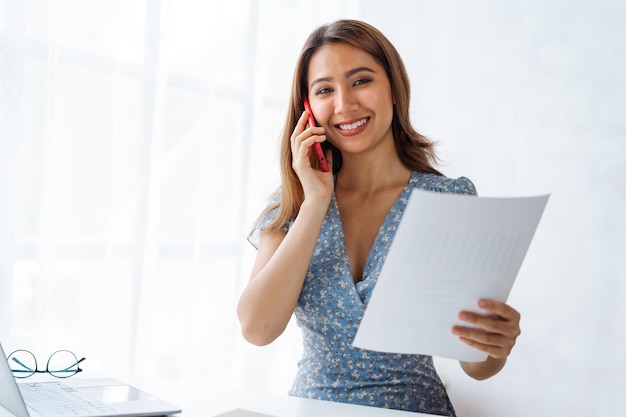 Young pretty Asian business woman talking on the phone when using laptop at working desk in modern office workplace