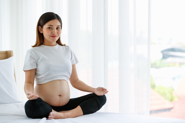 Young pretty Asian pregnant woman sitting on a bed doing exercise delightfully by Yoga. She is smiling and looking at a camera. Healthy happy mother concept.