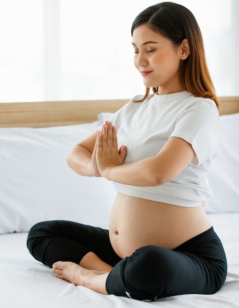 A young pretty Asian woman sitting on a bed joins her hands in front of her chest with a smiley face looking at a camera. Yoga is a great exercise for pregnant ladies.