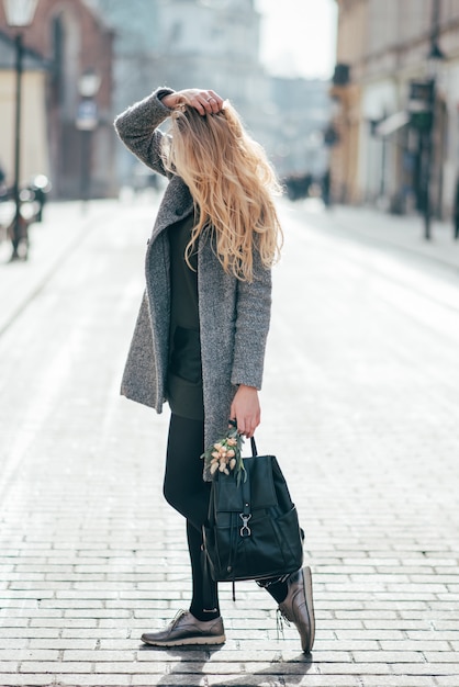 Young pretty blond woman with long blond wavy hair is in the street and holding her blond hair, the backpack and bouquet.