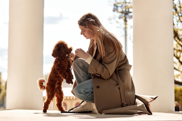 A young pretty blonde girl in a beige raincoat and jeans is squatting and training her cute pet red