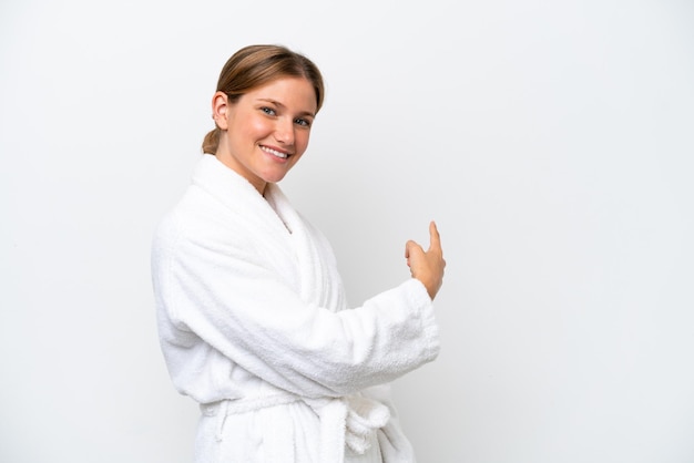 Young pretty blonde woman in bathrobe isolated on white background pointing back