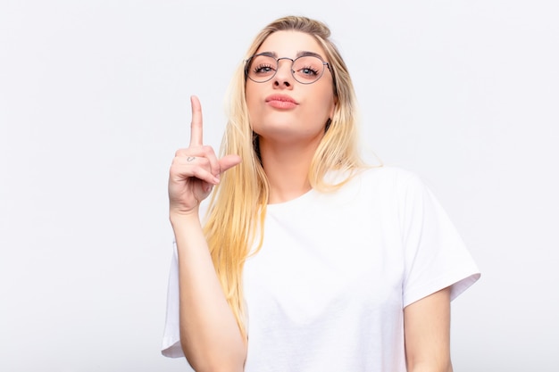 Young pretty blonde woman feeling like a genius holding finger proudly up in the air after realizing a great idea, saying eureka over white wall