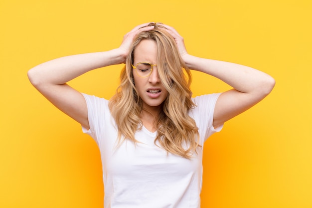 Young pretty blonde woman feeling stressed and frustrated, raising hands to head, feeling tired, unhappy and with migraine over color wall