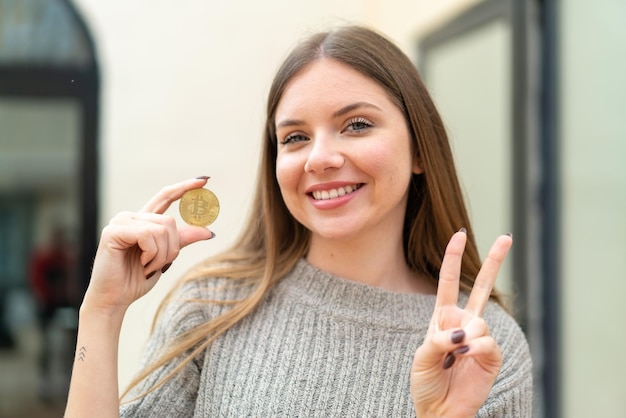 Young pretty blonde woman holding a Bitcoin at outdoors smiling and showing victory sign