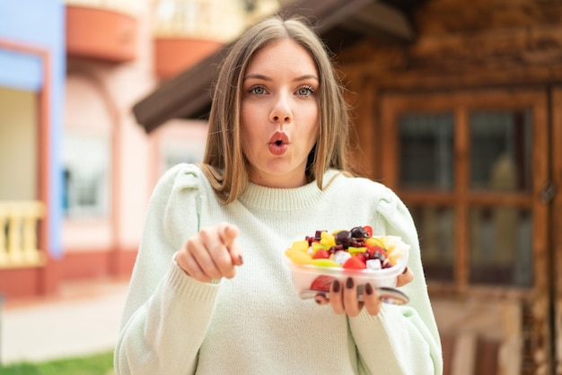 Young pretty blonde woman holding a bowl of fruit at outdoors surprised and pointing front