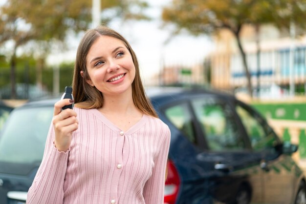 Young pretty blonde woman holding car keys at outdoors looking up while smiling