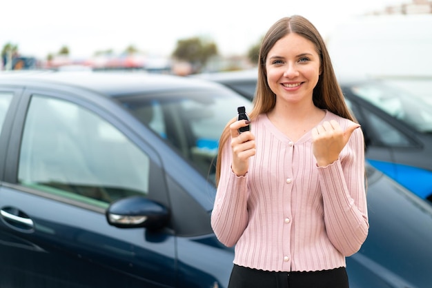 Young pretty blonde woman holding car keys at outdoors pointing to the side to present a product