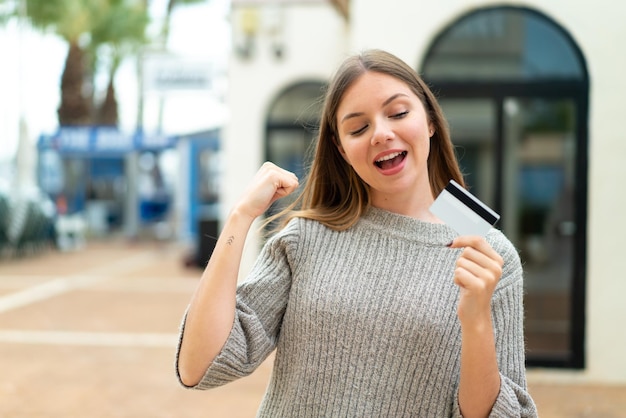 Young pretty blonde woman holding a credit card at outdoors celebrating a victory