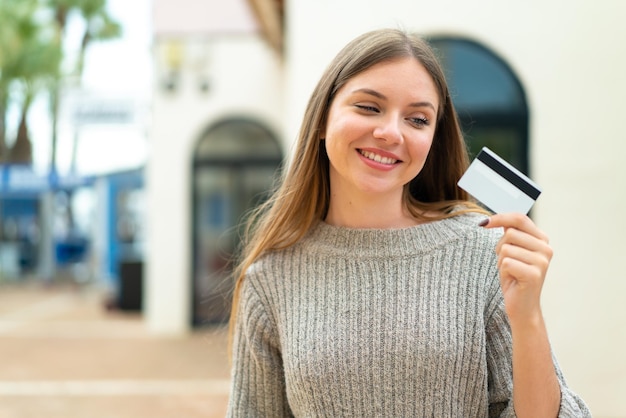 Young pretty blonde woman holding a credit card at outdoors with happy expression