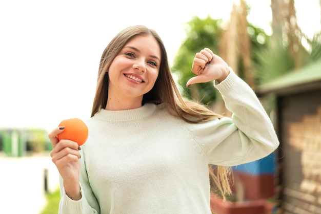 Young pretty blonde woman holding an orange at outdoors proud and selfsatisfied