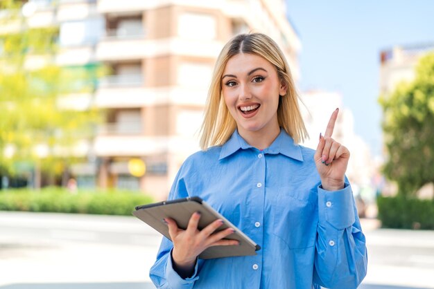 Young pretty blonde woman holding a tablet at outdoors pointing up a great idea