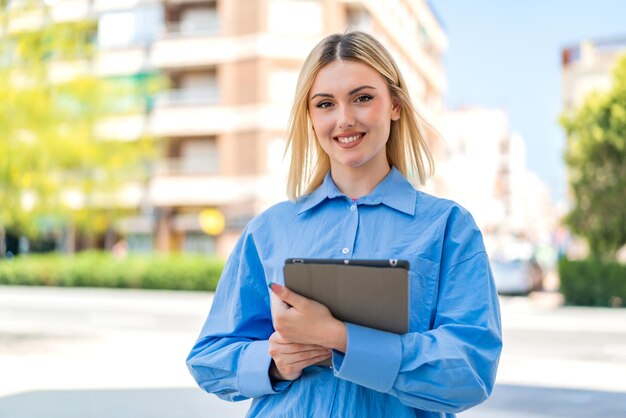 Young pretty blonde woman holding a tablet at outdoors smiling a lot