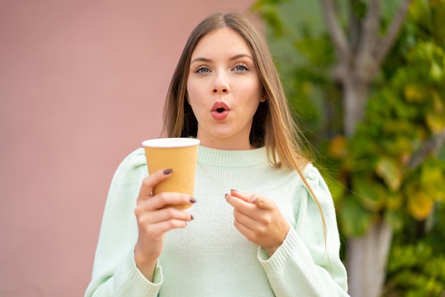 Young pretty blonde woman holding a take away coffee at outdoors surprised and pointing front