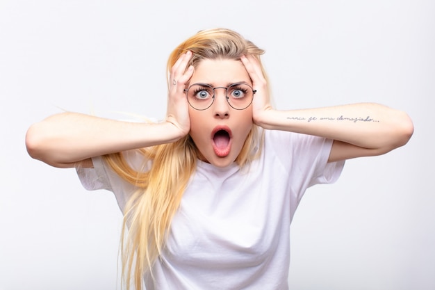 Young pretty blonde woman looking unpleasantly shocked with mouth wide open and covering both ears with hands against white wall