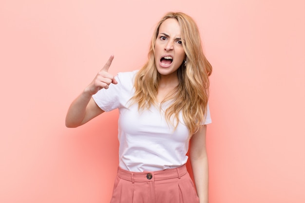 Young pretty blonde woman pointing at camera with an angry aggressive expression looking like a furious, crazy boss against flat color wall