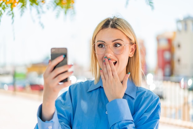 Young pretty blonde woman using mobile phone at outdoors with surprise and shocked facial expression