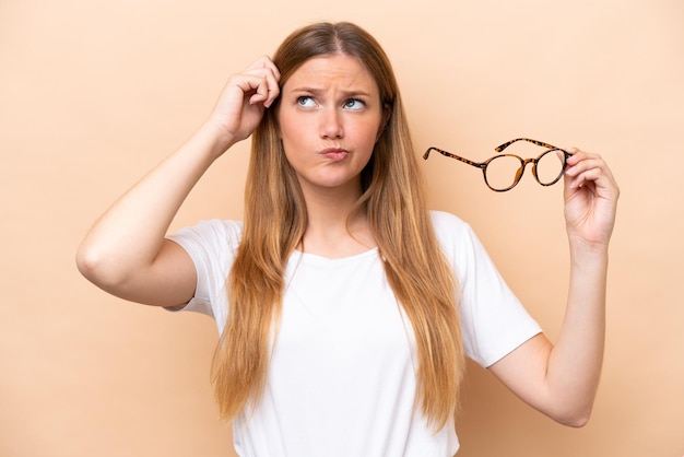 Young pretty blonde woman with glasses isolated on beige background having doubts and with confuse face expression