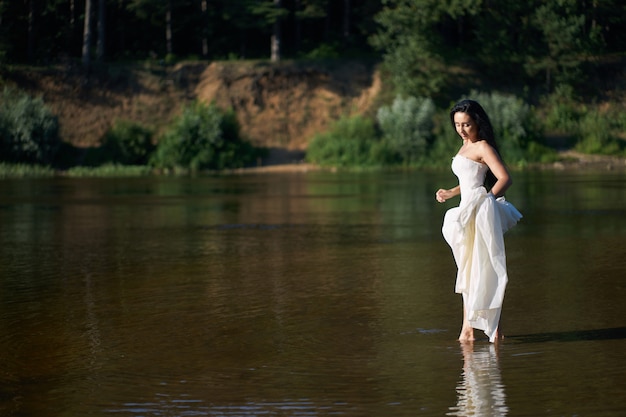 Young pretty brunette woman in white wedding dress walking on the river barefoot