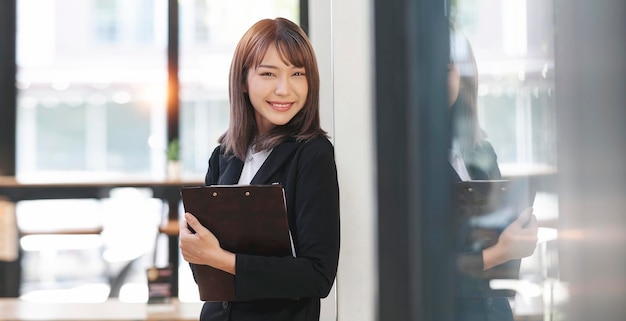 Young pretty businesswoman holding file folder standing in office smiling and looking at camera