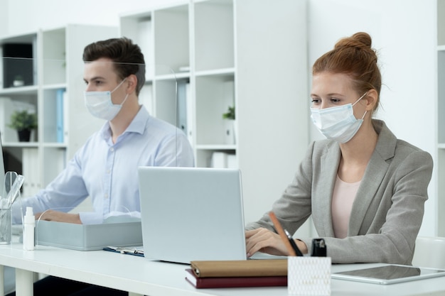 Young pretty businesswoman in protective mask and grey suit looking at laptop display while analyzing or entering data against co-worker