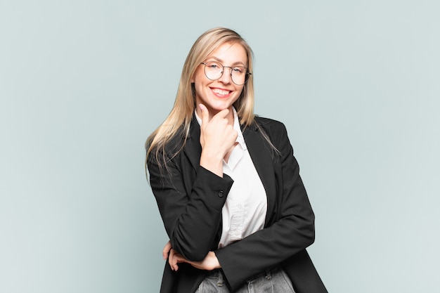 Young pretty businesswoman smiling, enjoying life, feeling happy, friendly, satisfied and carefree with hand on chin