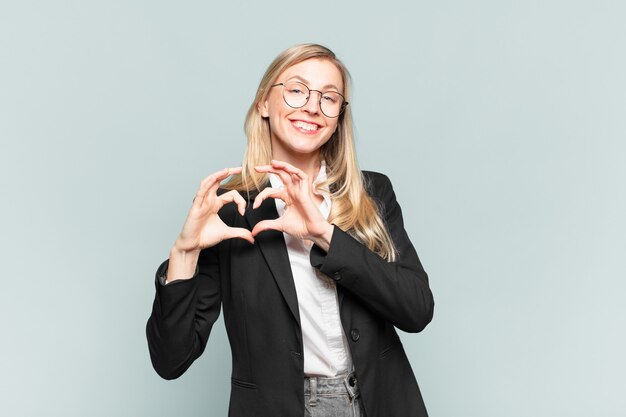 Young pretty businesswoman smiling and feeling happy, cute, romantic and in love, making heart shape with both hands