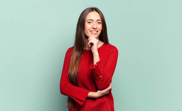 Young pretty casual woman looking happy and smiling with hand on chin, wondering or asking a question, comparing options